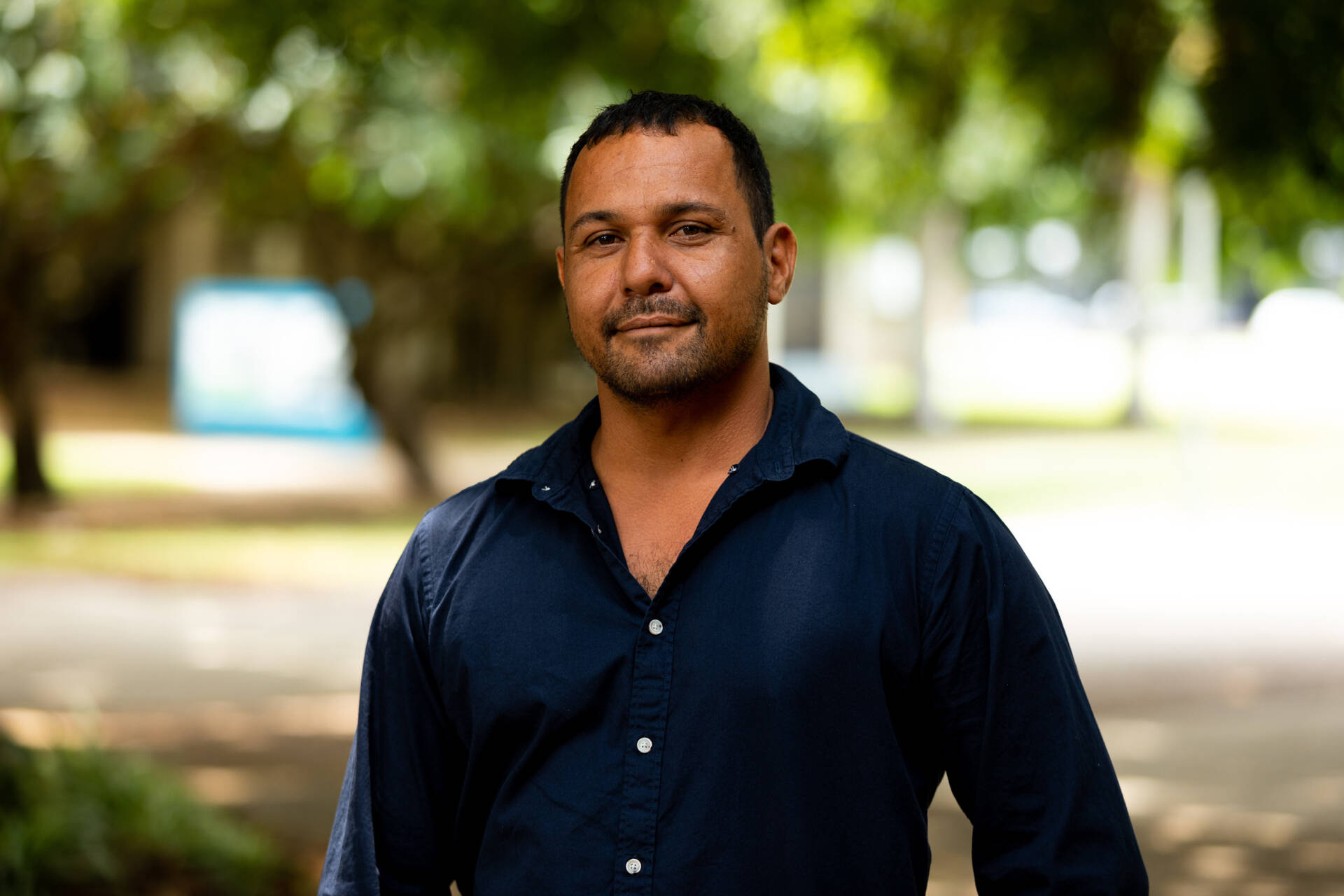 A young Aboriginal man in a blue button-up shirt smiles for the camera while standing outside.