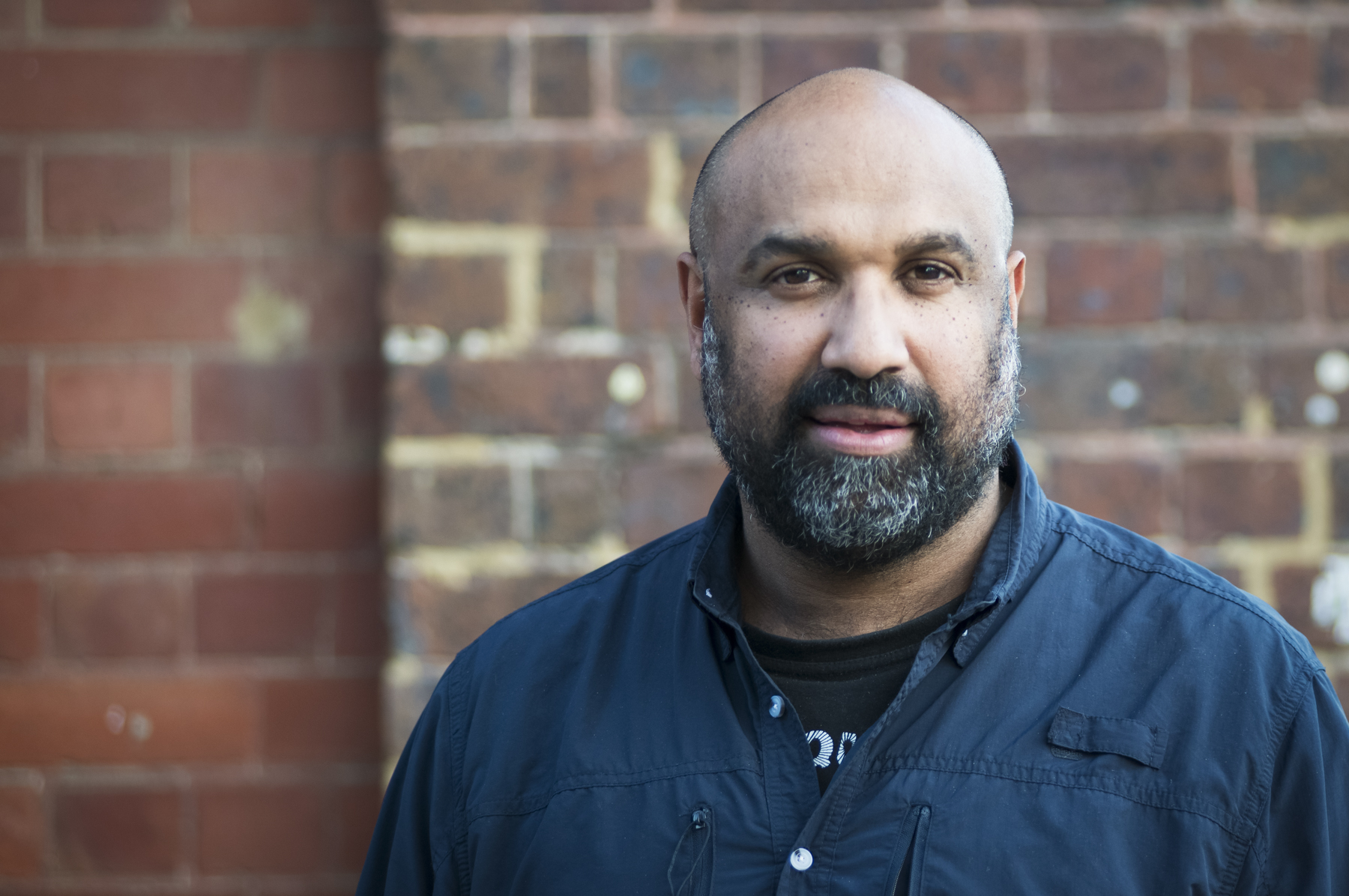 A middle-aged, bald Indigenous man in a blue shirt looks at the camera in front of a brick wall.